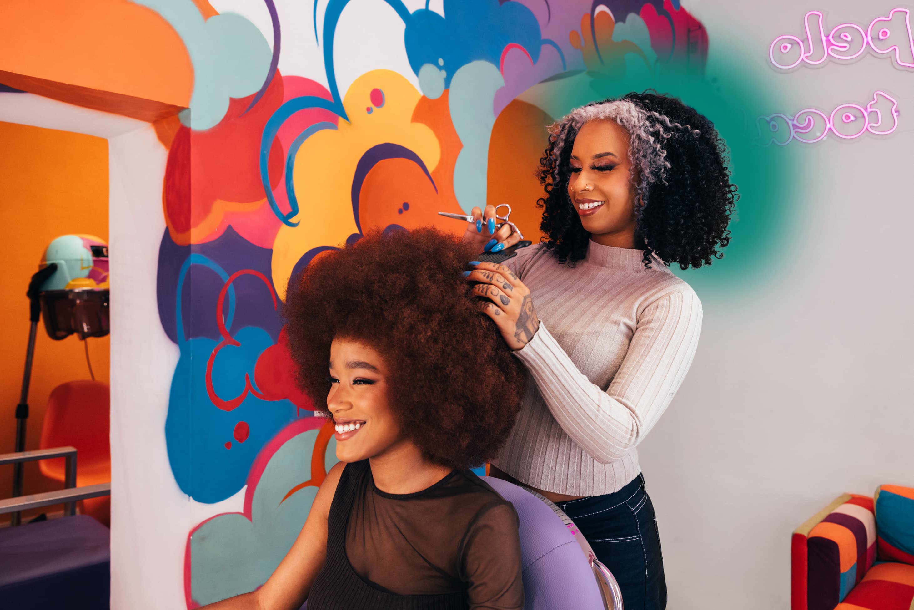 Hairstylist styling a smiling client's hair in a colourful mural-painted salon